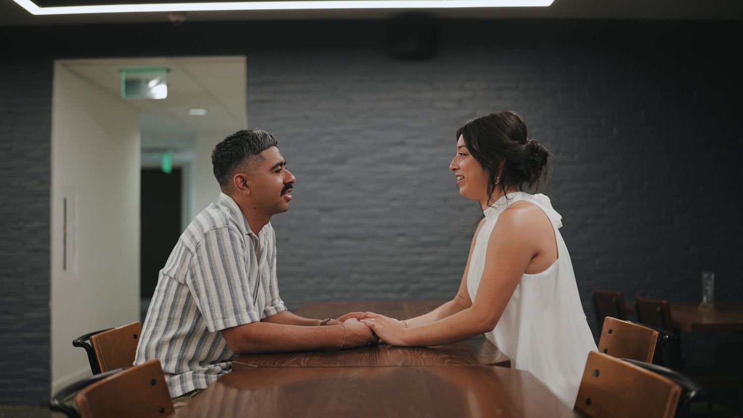 Two people sitting at a wooden table having an intimate conversation in a modern interior setting.
