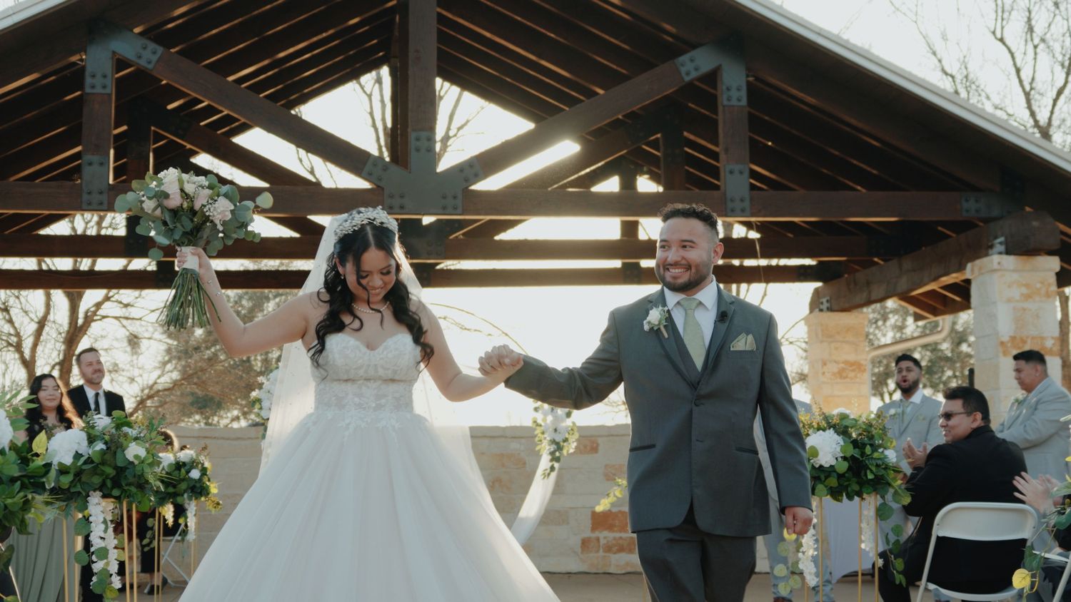 Newlyweds celebrate under a rustic wooden pavilion decorated with flowers.