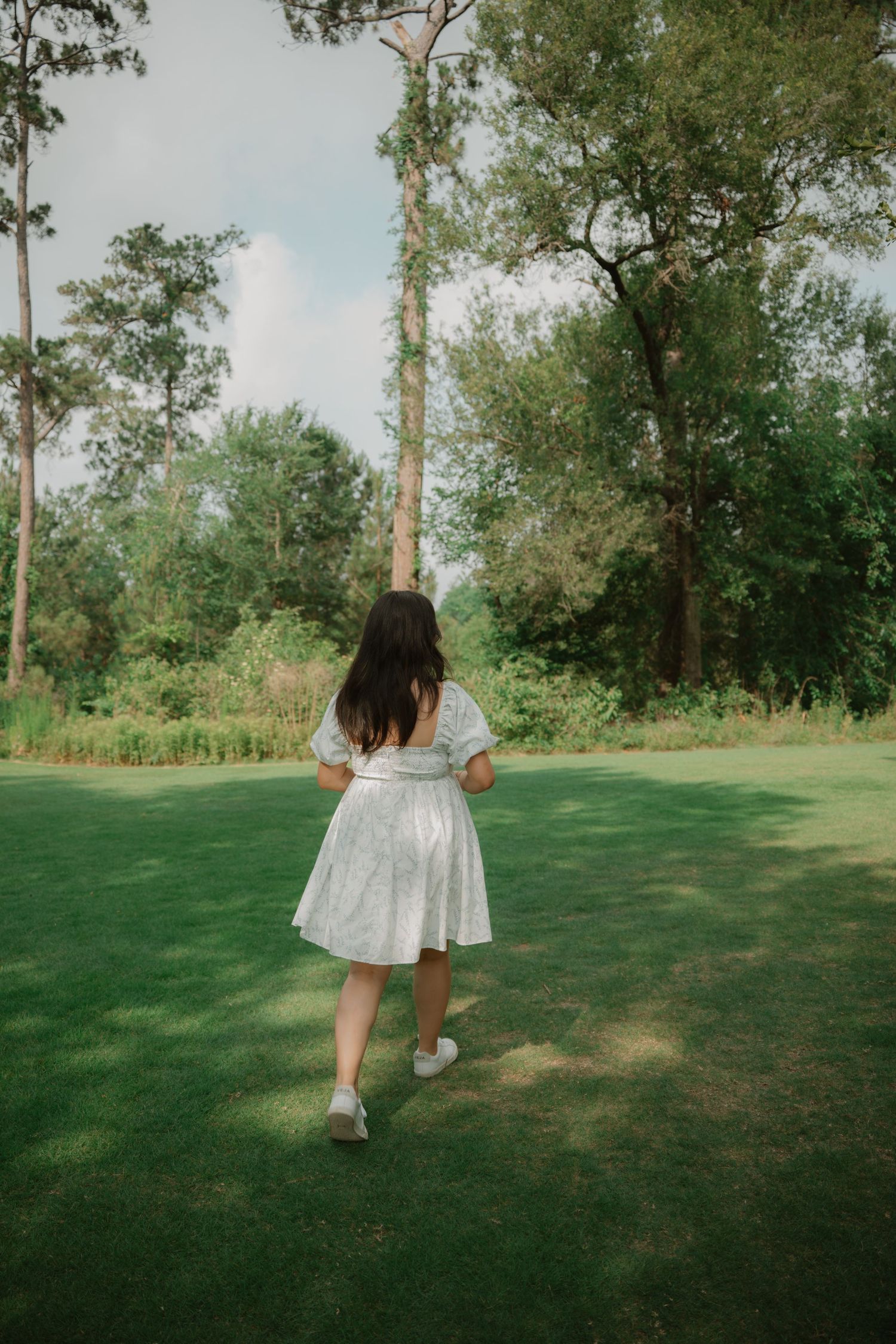 A person in a white dress spins playfully on a grassy lawn surrounded by trees.