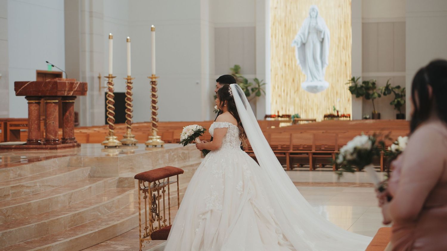 A bride in a flowing white gown and veil stands at the altar of a Catholic church with illuminated Marian statue.
