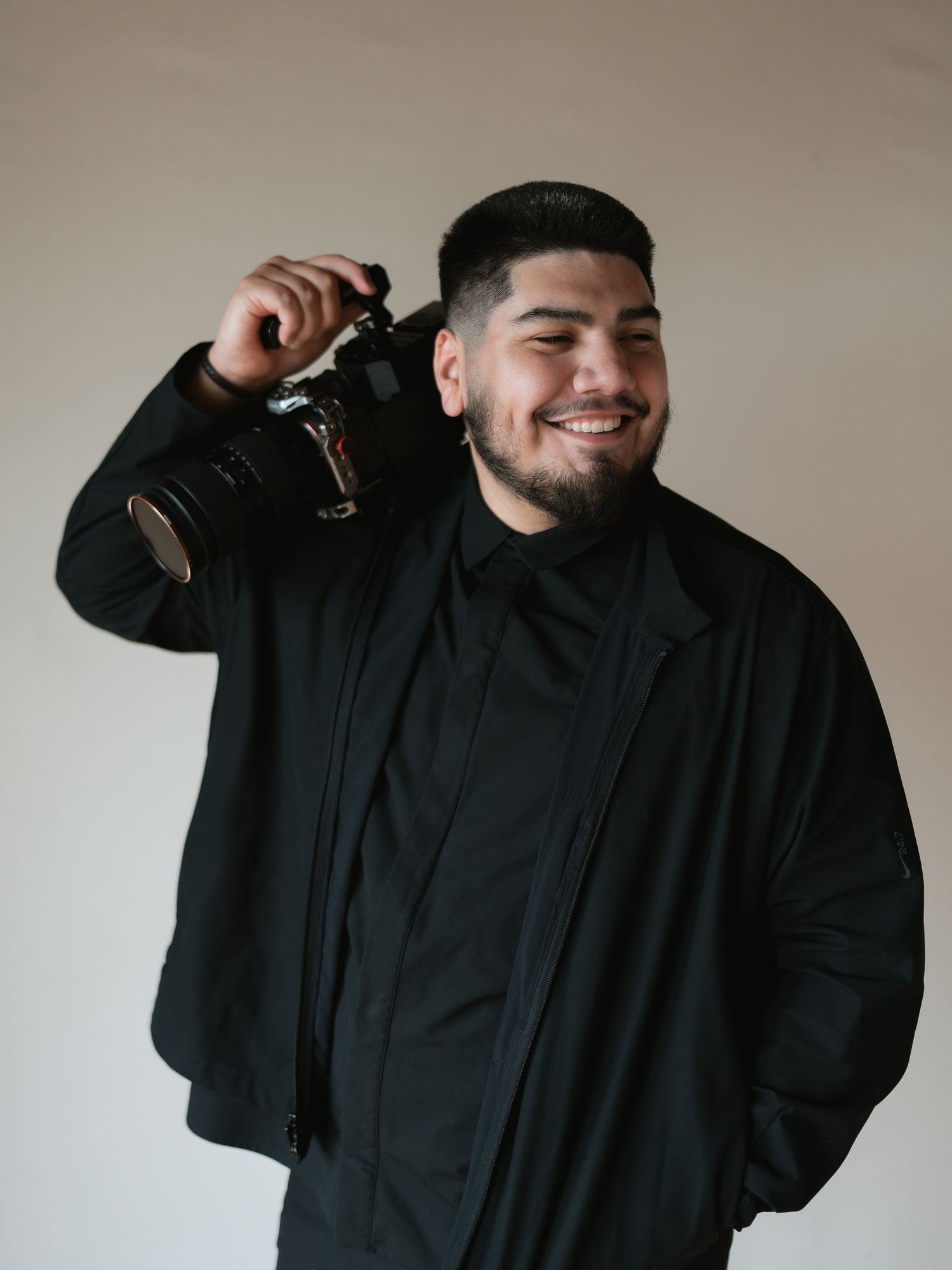 Smiling young man in all-black outfit holding a professional camera on his shoulder against a light background.