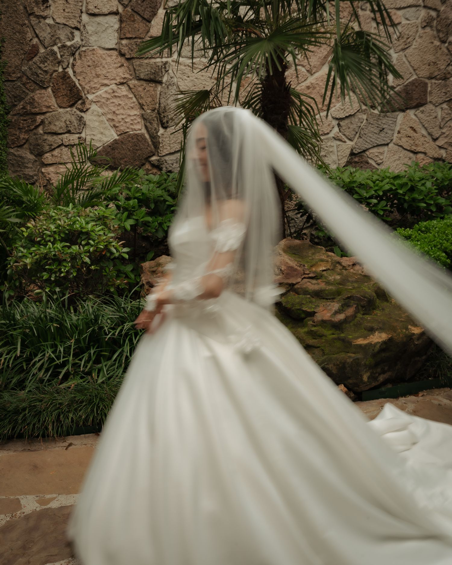 Artistic motion blur photograph of a flowing white wedding gown against a stone wall with tropical plants.