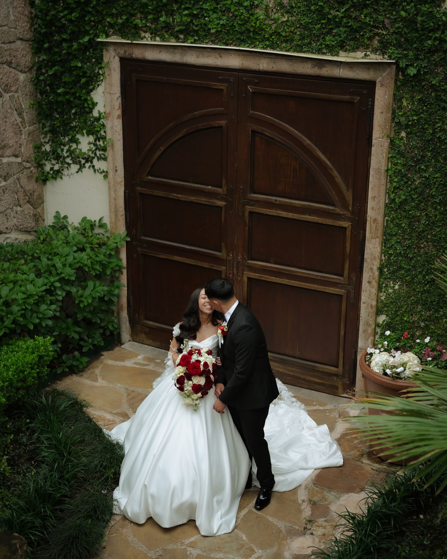 Wedding couple standing by a large wooden door surrounded by ivy and tropical landscaping.