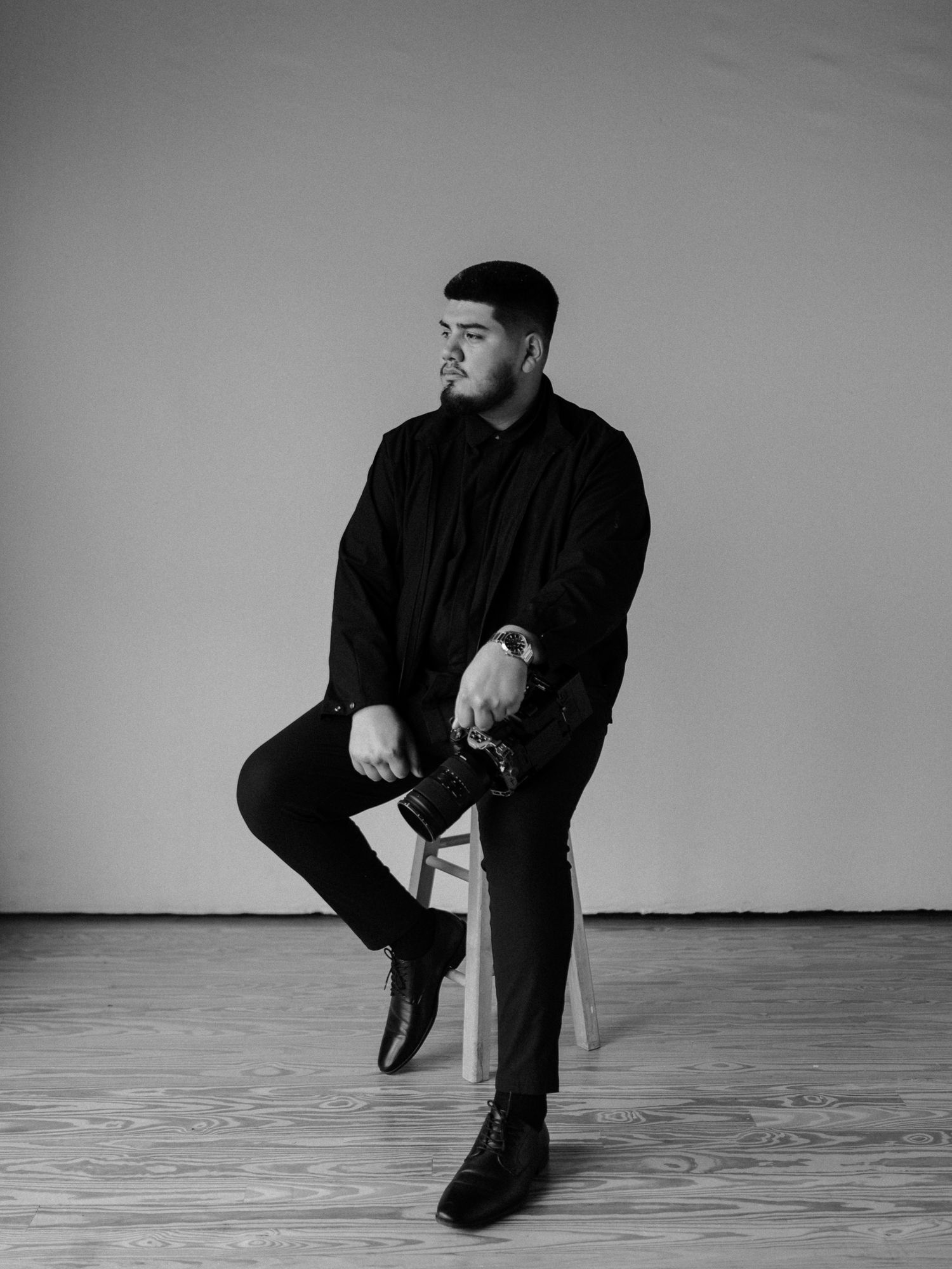 Black and white portrait of a man in all-black outfit sitting on a stool holding a camera in a studio.