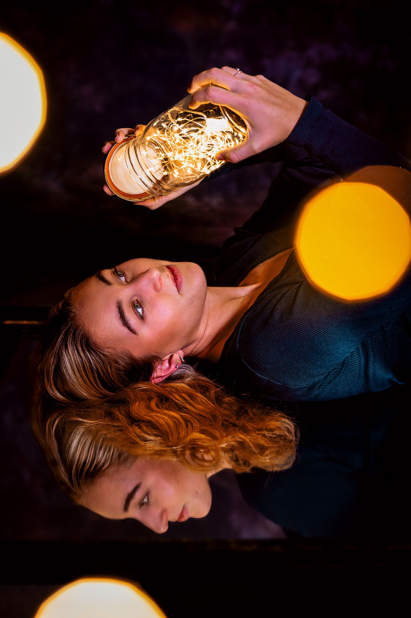 Woman holding a glowing mason jar with fairy lights, reflected on a mirror surface with bokeh light effects.