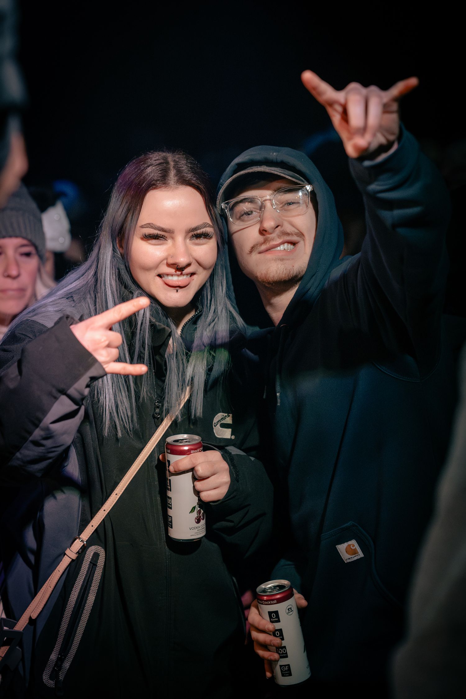 Concert attendees in dark clothing holding beverages and making peace signs at a nighttime event.