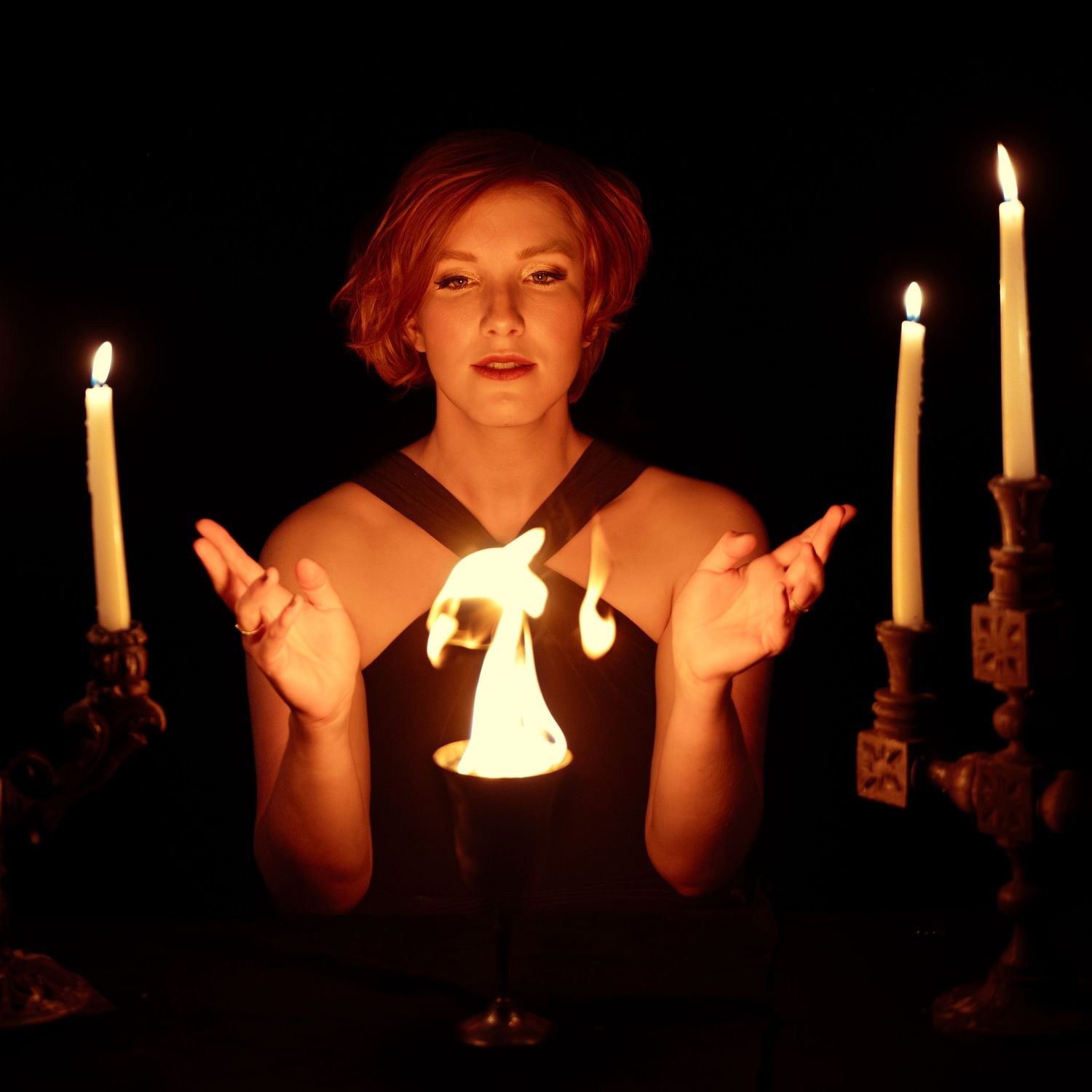 Woman in mystical setting with candles performing a ritual gesture with glowing light between her hands in dark atmosphere.