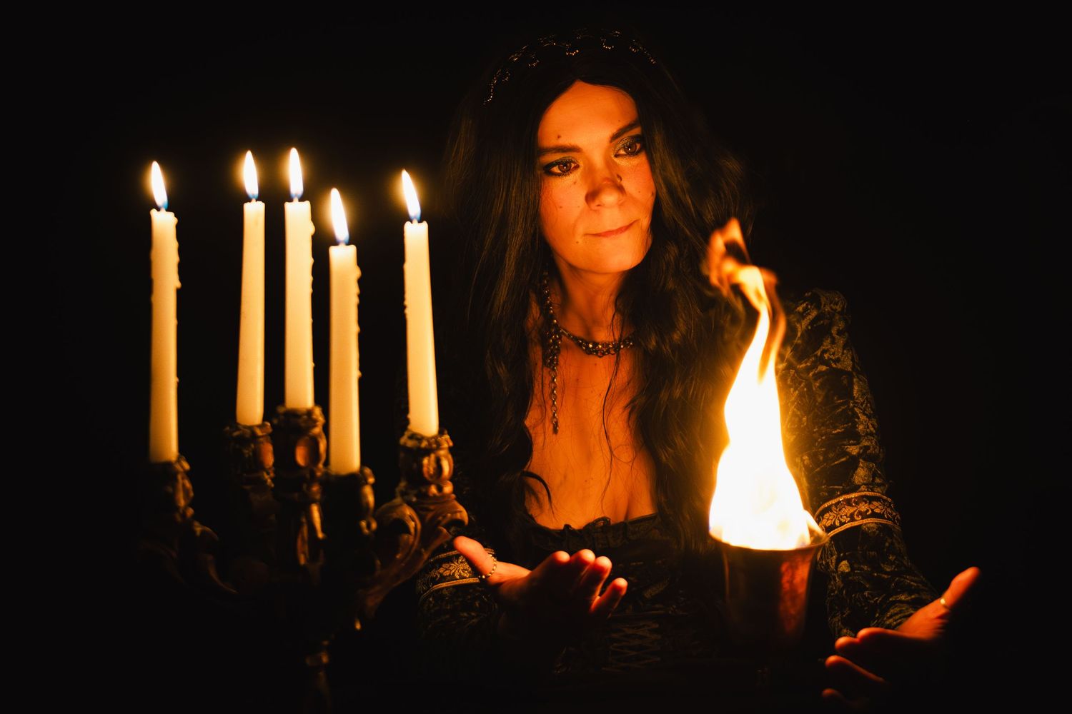 Woman in dark gothic attire holds flame in palm beside ornate candelabra with lit white candles in dramatic lighting.