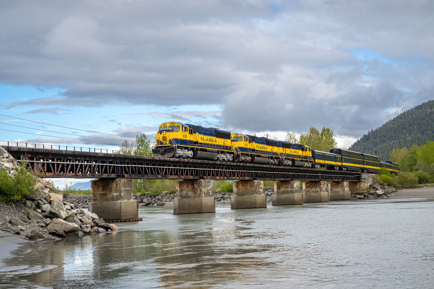 Yellow freight train crosses river bridge with mountain landscape in background. Alaska Railroad Coastal Classic.