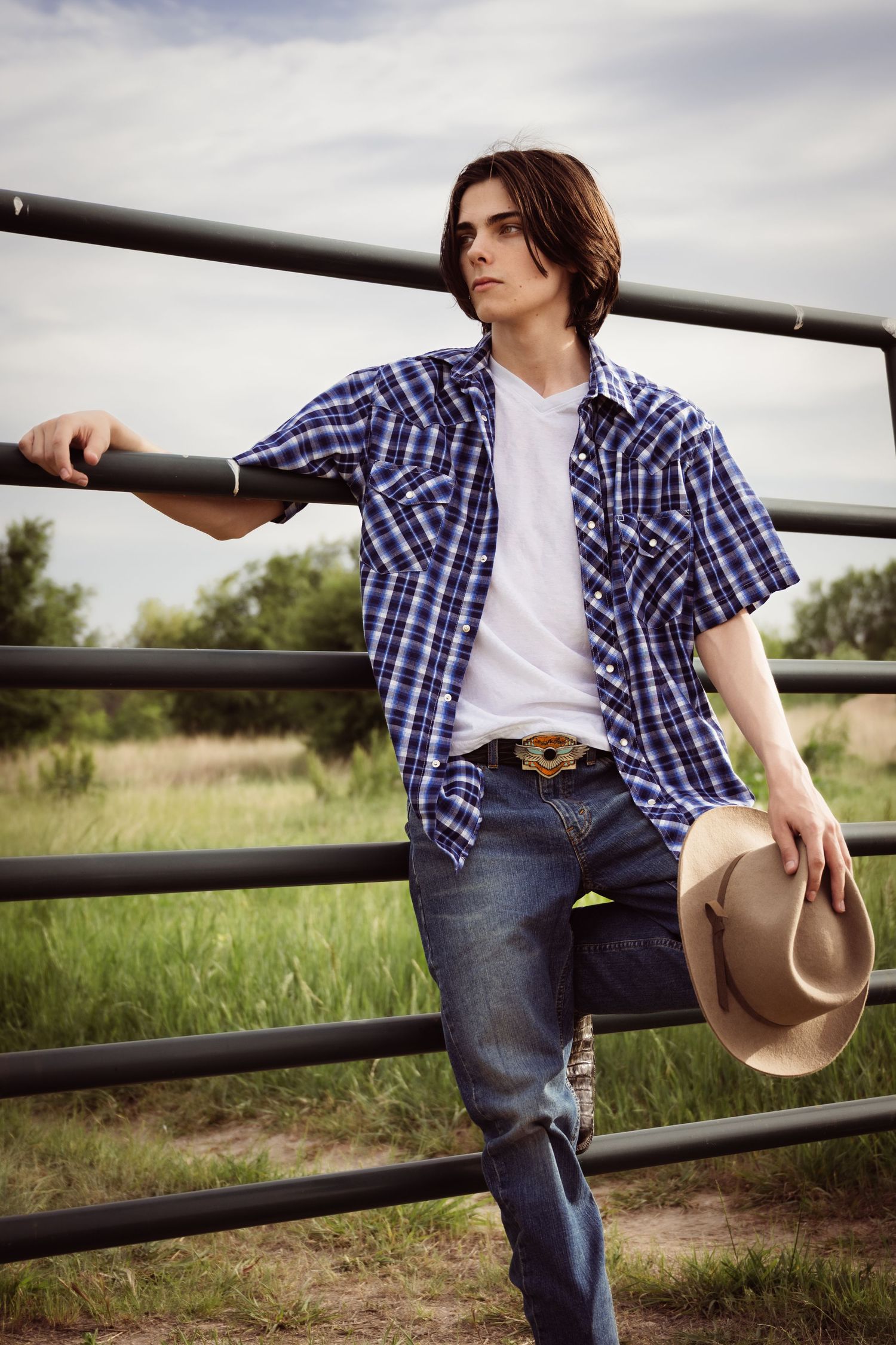 Western-style portrait showing someone in plaid and denim leaning against a ranch fence.