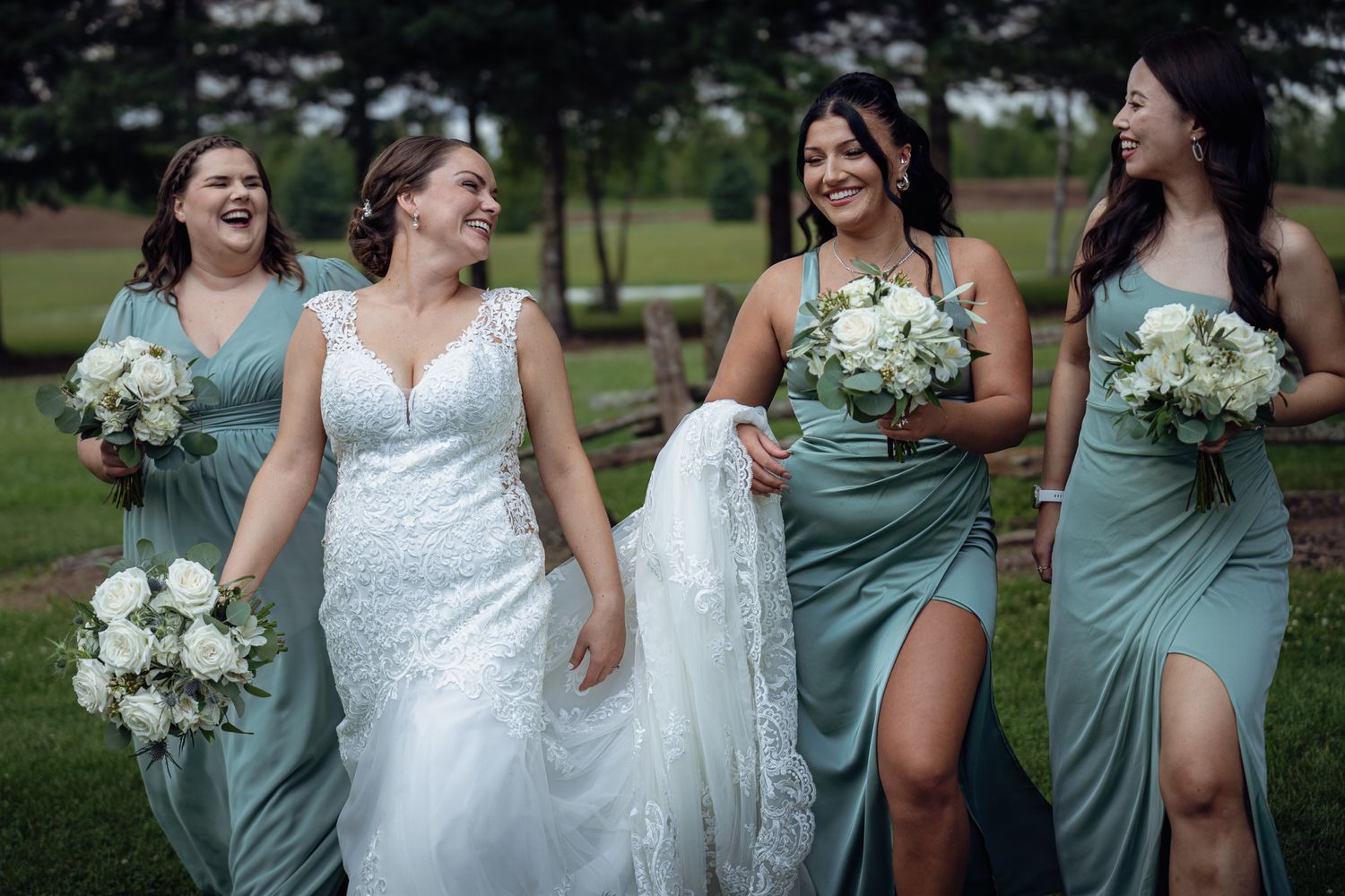 Bride in white lace gown laughing with three bridesmaids in sage green dresses holding white bouquets outdoors.