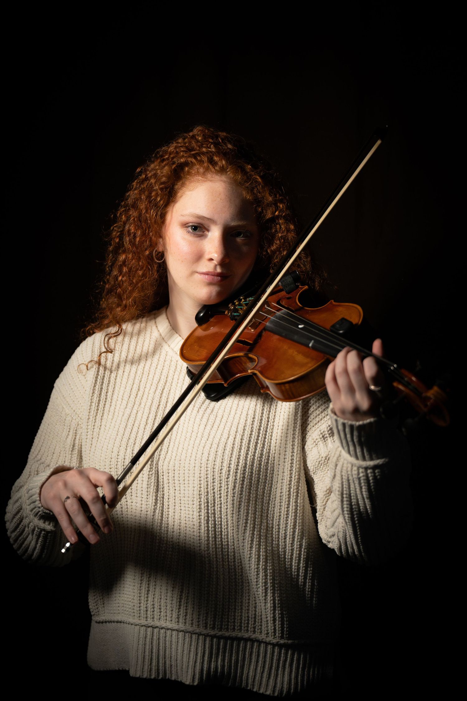 Young woman with curly red hair playing violin against dark background wearing cream sweater.