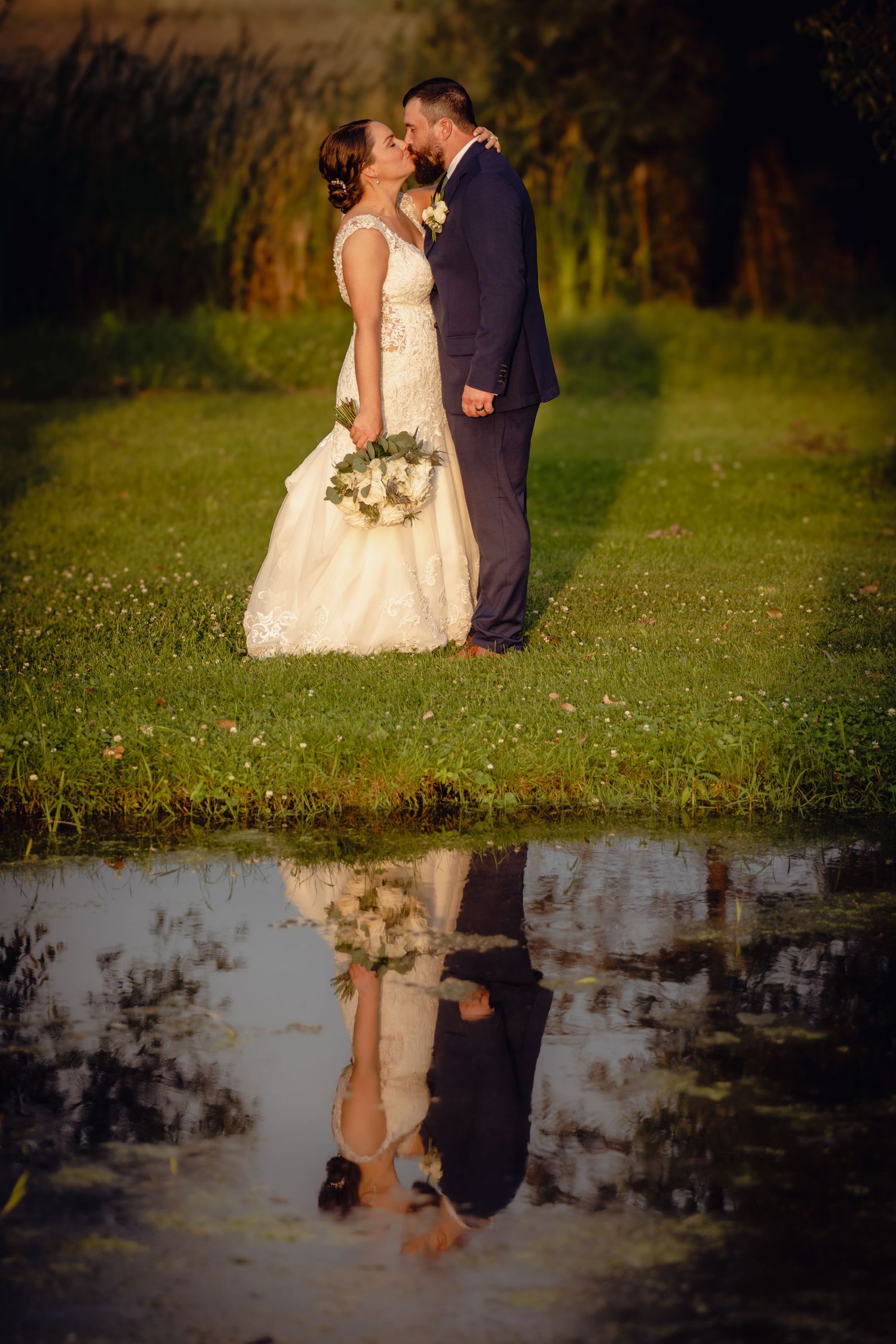 Wedding couple standing in a field with their reflection visible in a puddle at sunset.