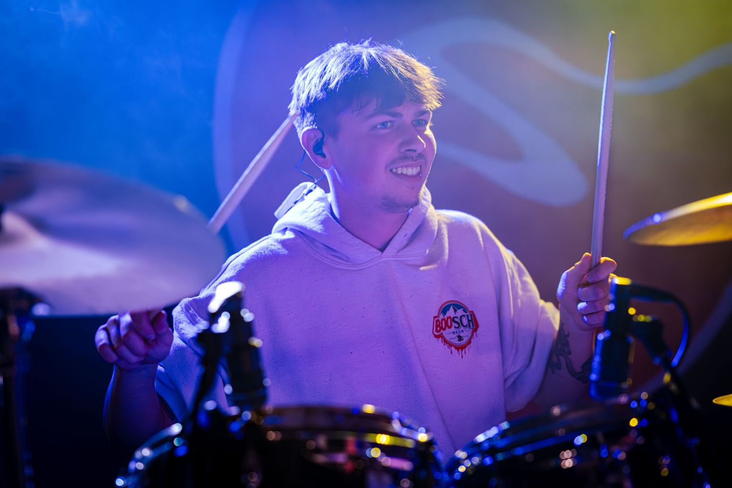 Concert lighting illuminates a drummer performing on stage with colorful blue backlighting.
