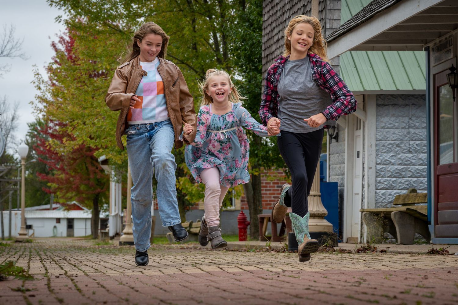 Three girls joyfully running hand in hand on brick path outdoors among autumn trees.