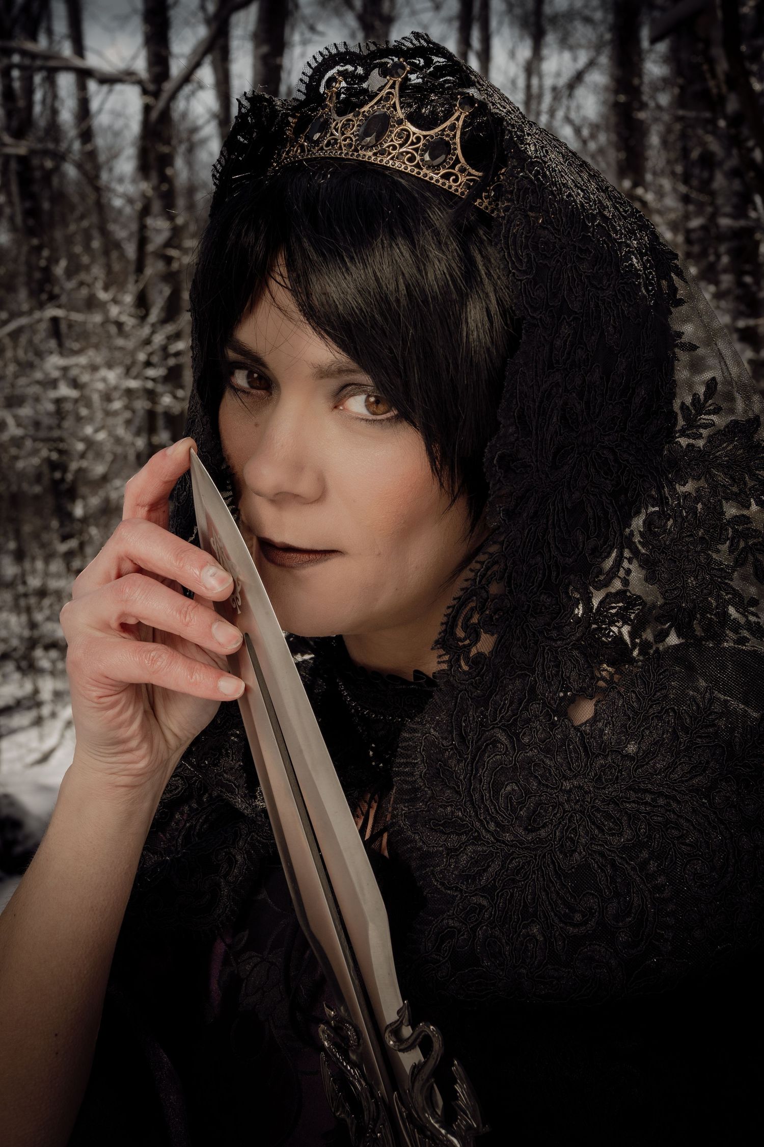 Woman in black lace hood and crown holding a sword blade in a snowy winter forest, gothic fantasy cosplay portrait.