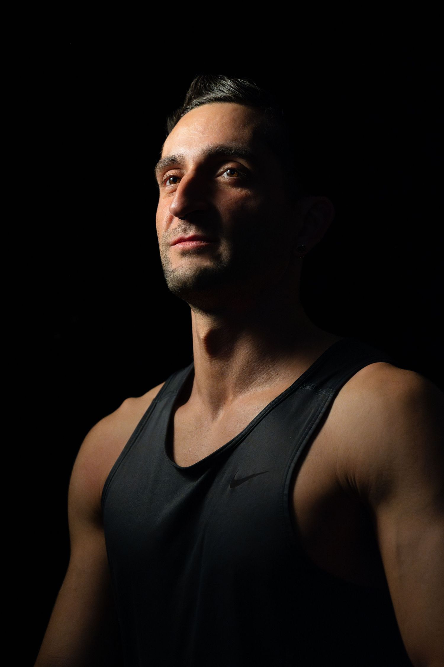 A muscular man in a black Nike tank top poses against a dark background with dramatic studio lighting.