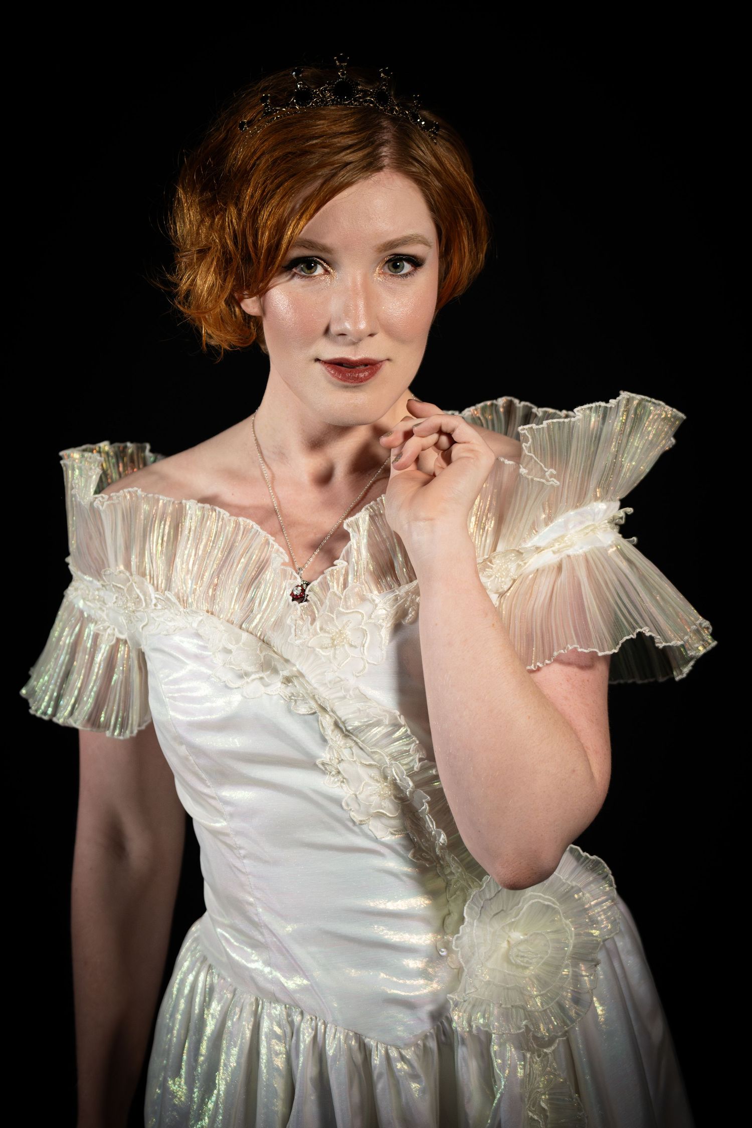 Red-haired woman in white iridescent ruffled gown wearing tiara poses against black background.