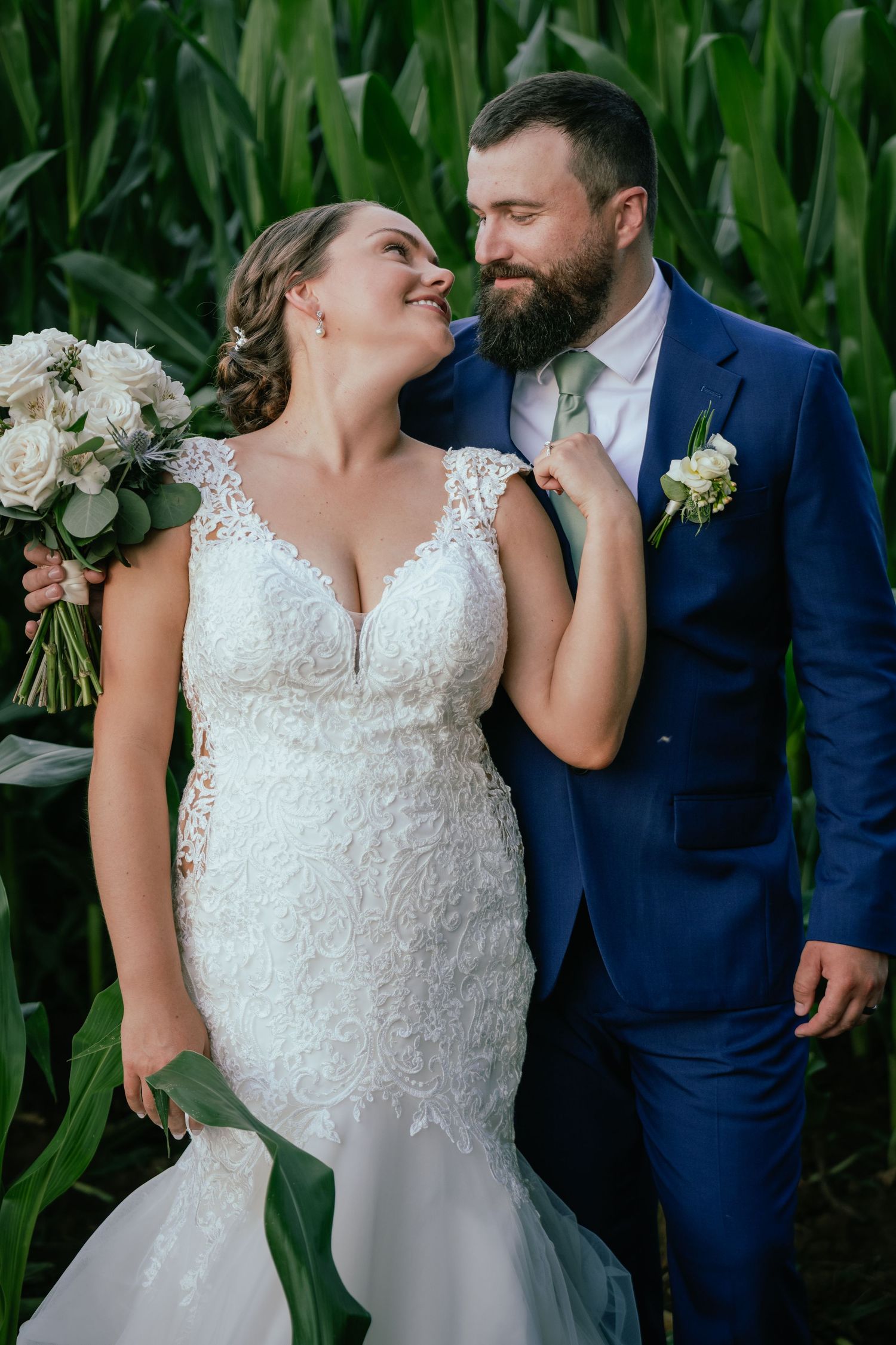 Bride in lace mermaid gown and bearded groom in navy suit share a romantic moment in a cornfield.