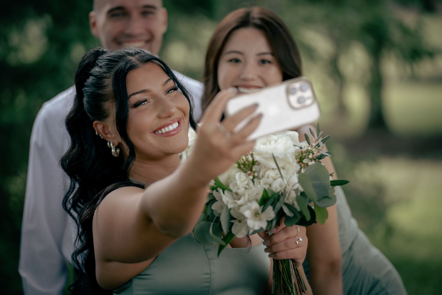 Bride smiling while taking a selfie with a bridesmaid, holding a white floral bouquet outdoors.