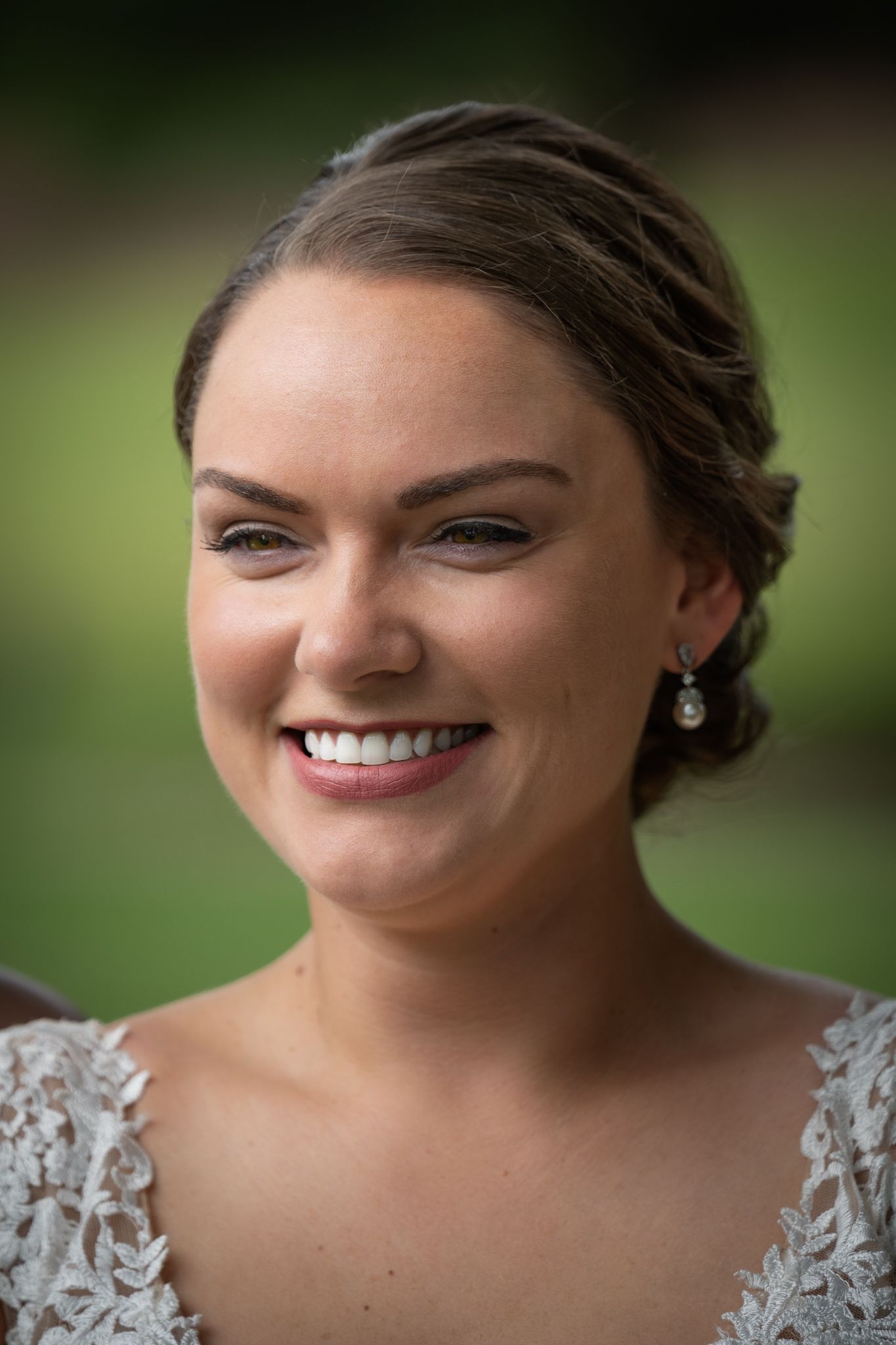 Smiling bride with updo hairstyle and pearl earrings wearing lace wedding dress outdoors.