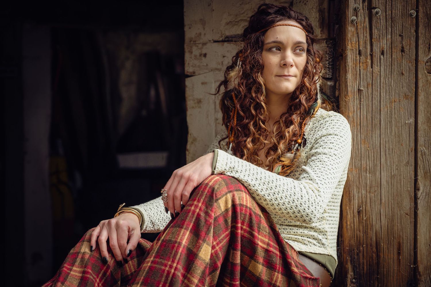 Curly-haired woman in plaid pants and headband sits relaxed against a rustic wooden wall.