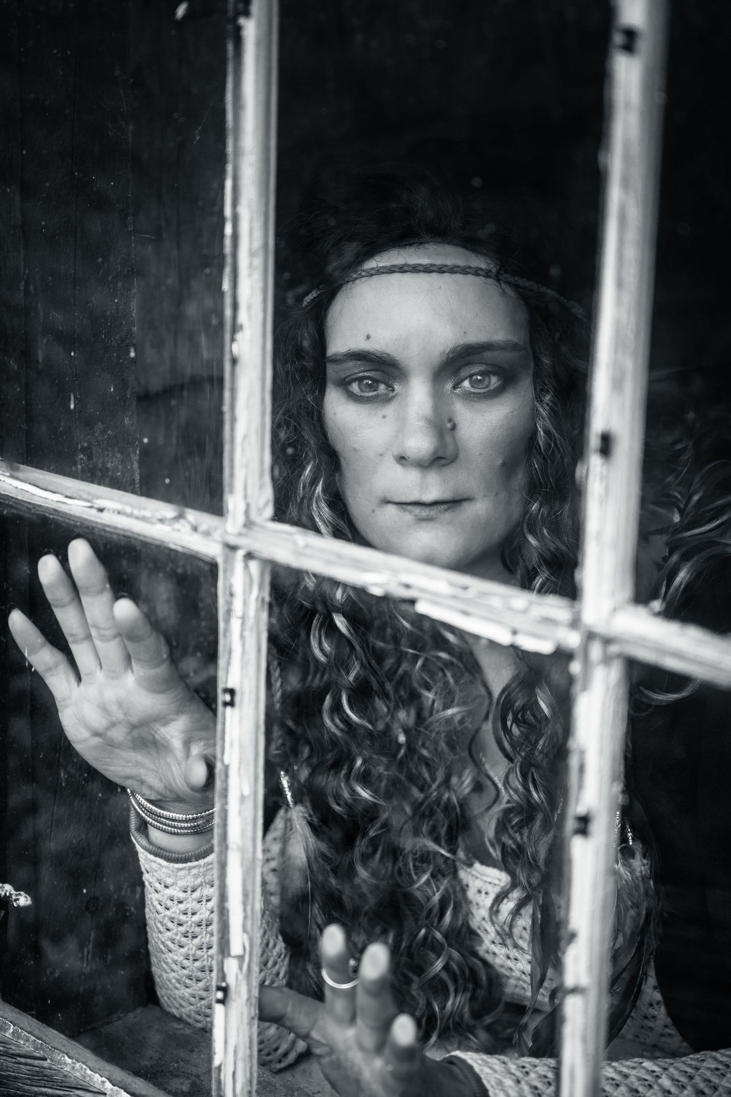 Black and white portrait of a curly-haired woman pressing her hand against an old window.