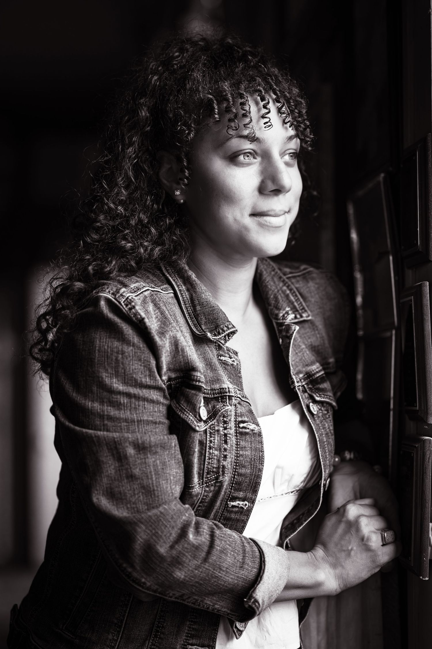 Artistic black and white portrait showing curly hair and denim jacket in dramatic lighting.