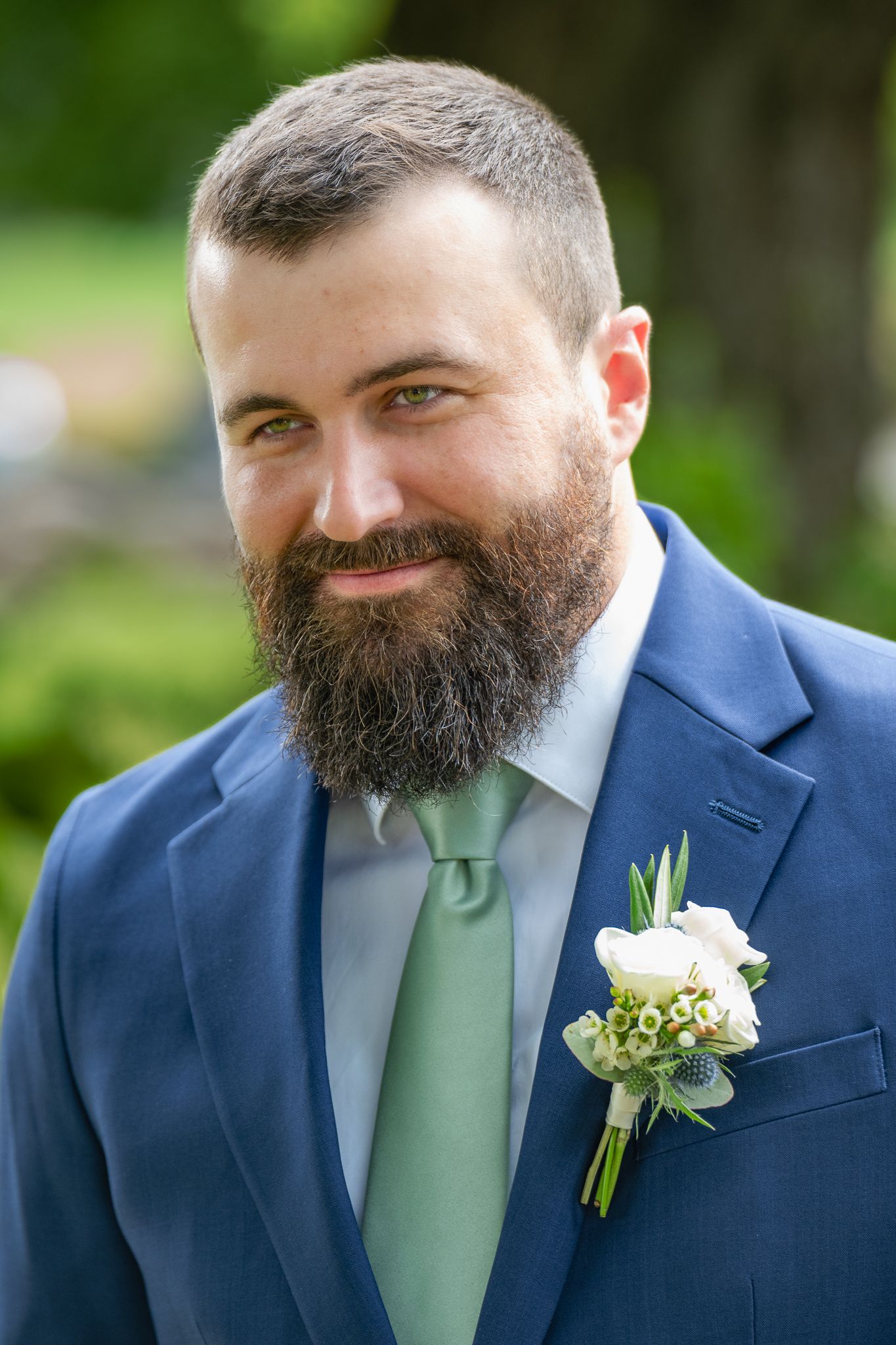 Bearded groom in navy blue suit with sage green tie and white boutonniere smiles outdoors at wedding.