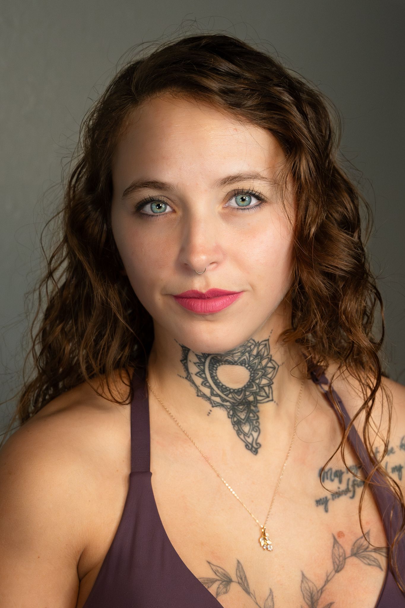 Young woman with curly brown hair, blue eyes, neck tattoo, and floral chest tattoos wearing a purple top.
