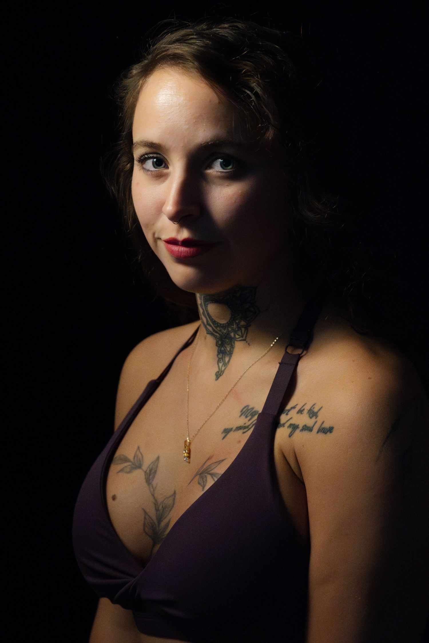 Young woman with tattoos on neck and chest wearing a purple bikini top poses against a dark background.