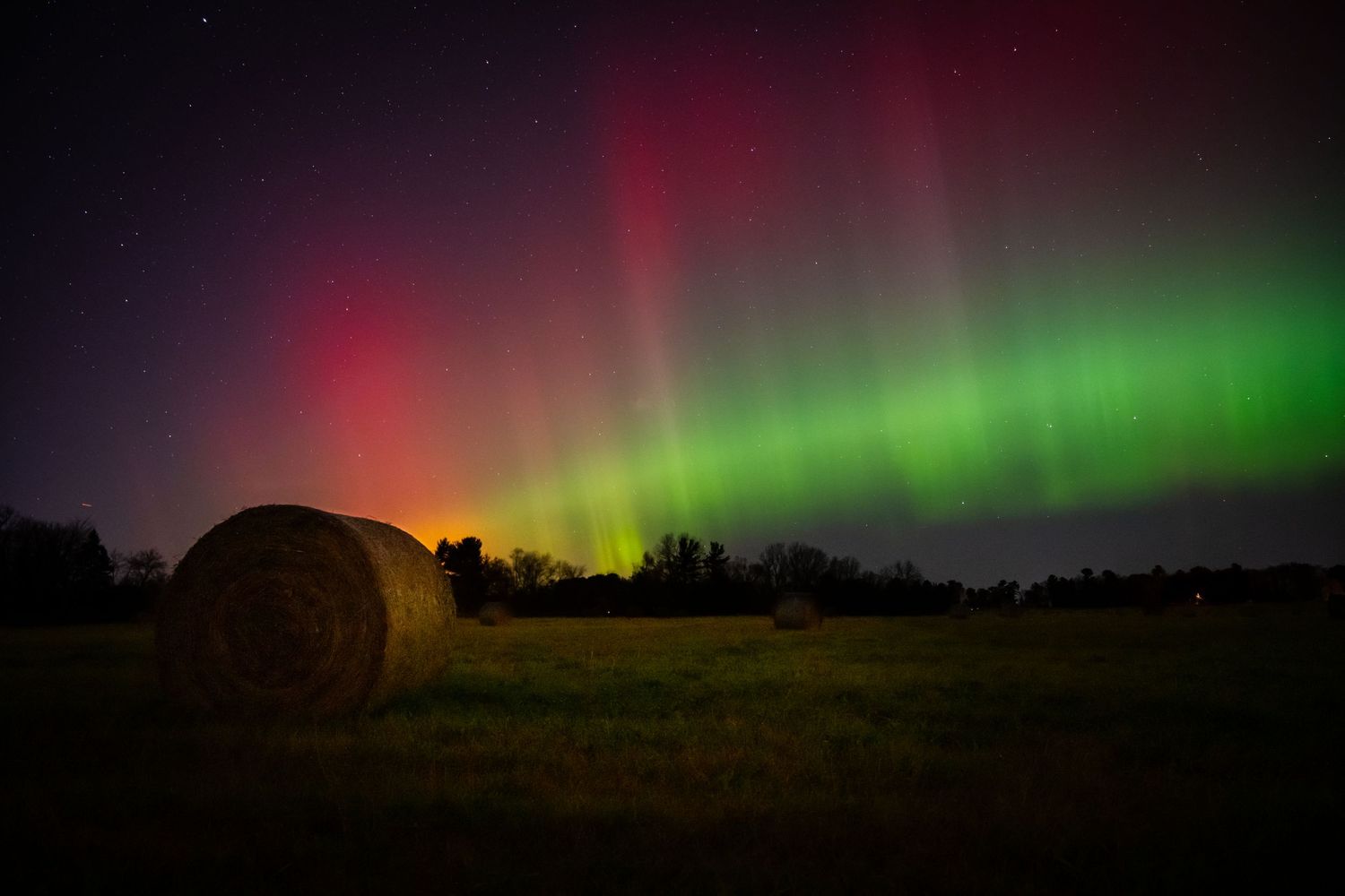 Stunning green and red aurora borealis lights dance across the dark night sky over a rural landscape.
