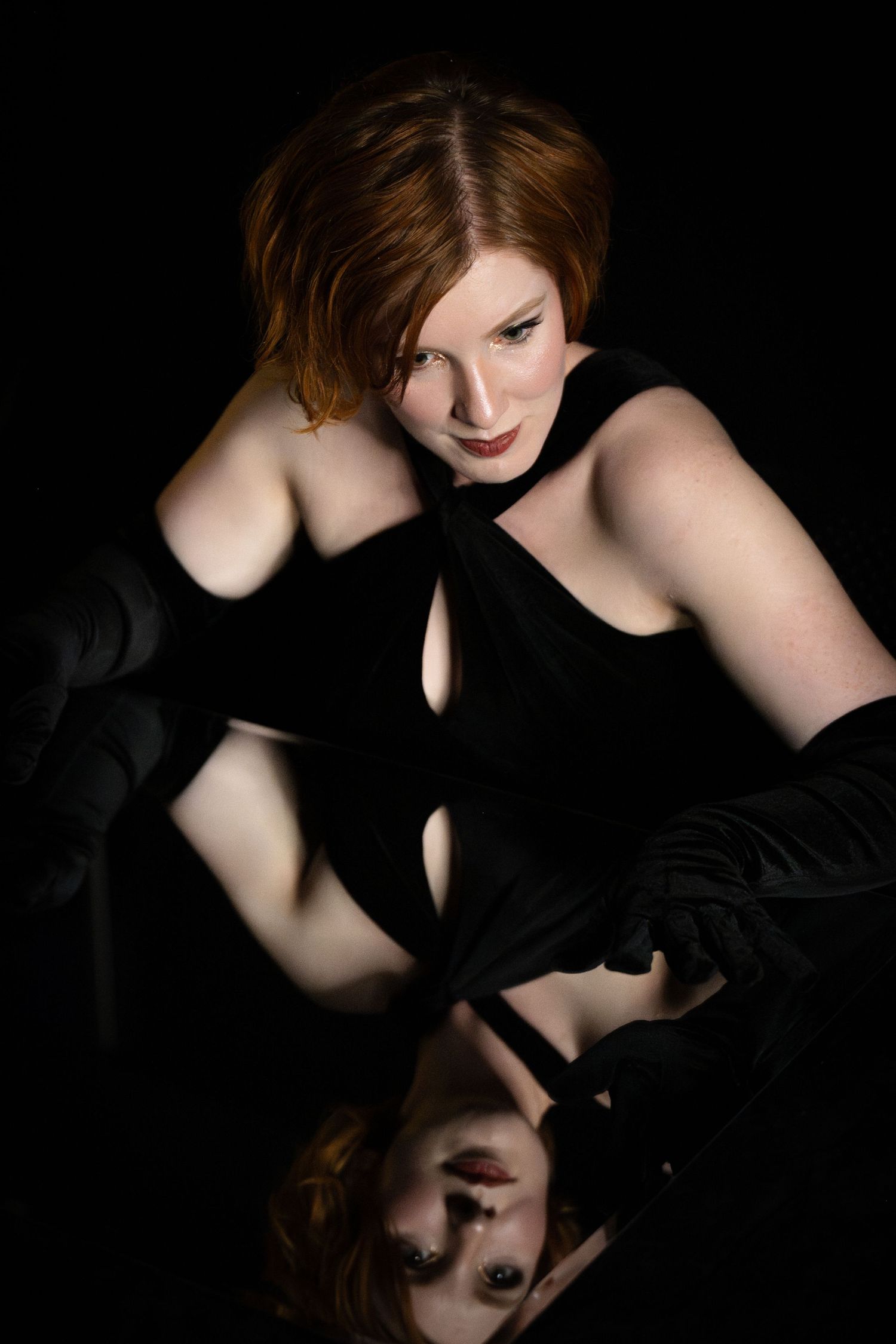 Elegant redhead woman in black dress and gloves poses with mirror reflection on dark background.