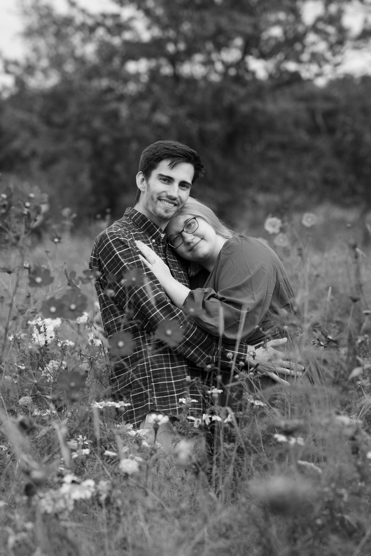 Black and white photo of a couple embracing in a wildflower field, smiling warmly.