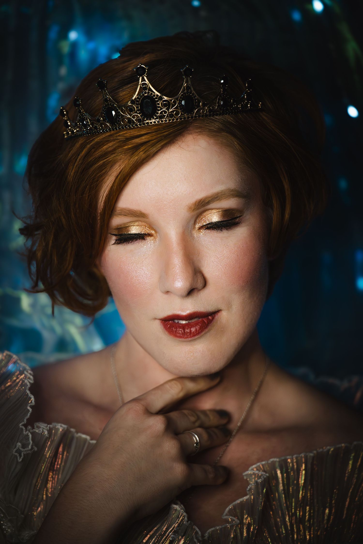 A woman wearing a gold crown and elegant dress poses with eyes downcast in a fantasy portrait.