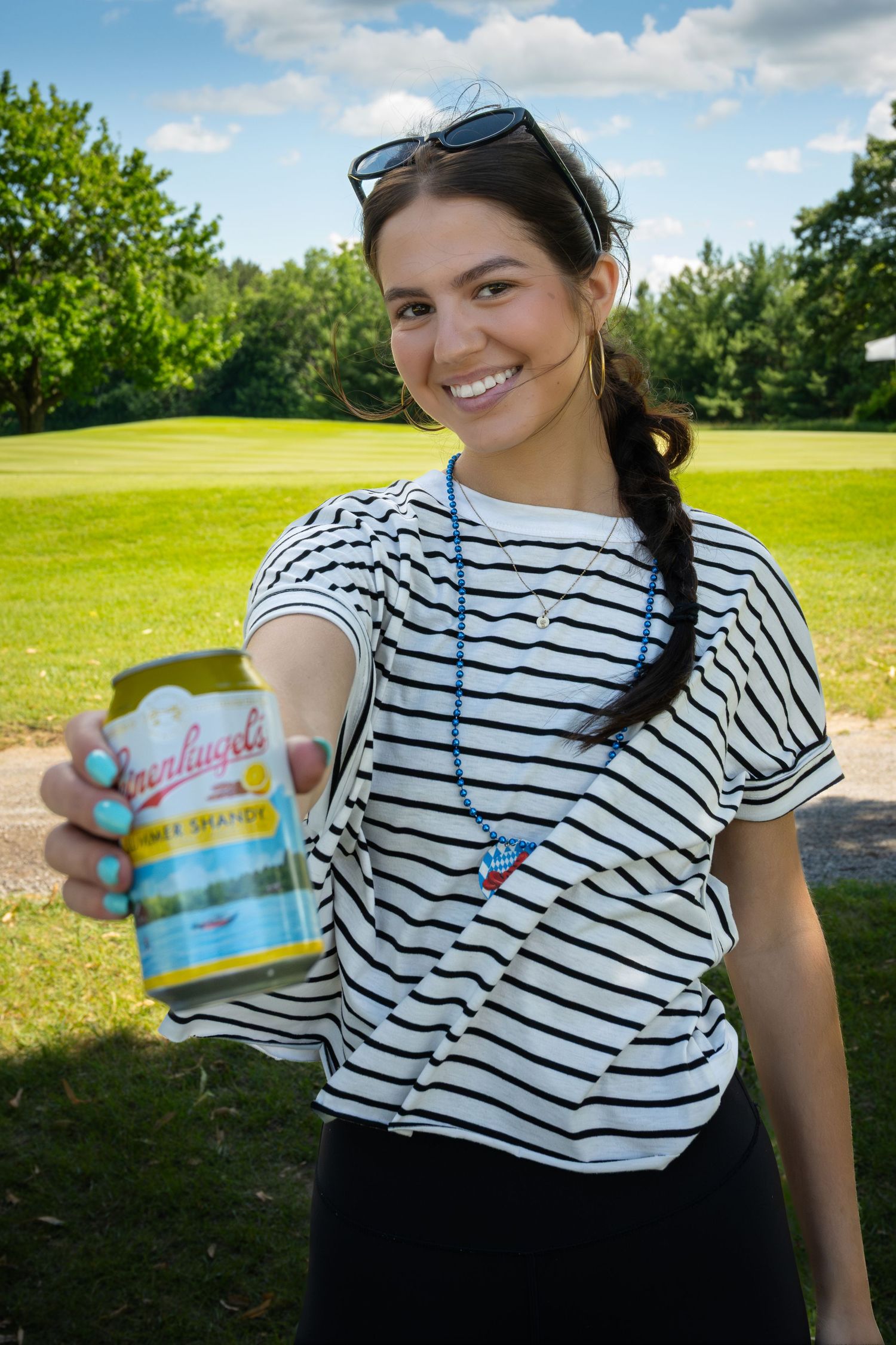 Smiling woman holding out a can of Greenlandic lemonade outdoors on a sunny golf course.