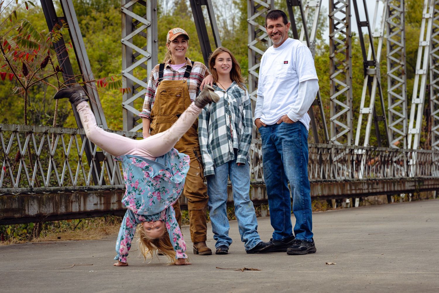Family of four poses on iron bridge with child doing a cartwheel in autumn setting.