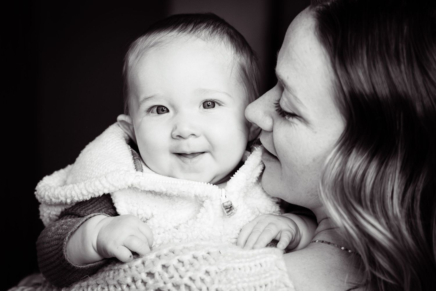 Black and white photo of a smiling baby being kissed on the cheek by a loving mother.
