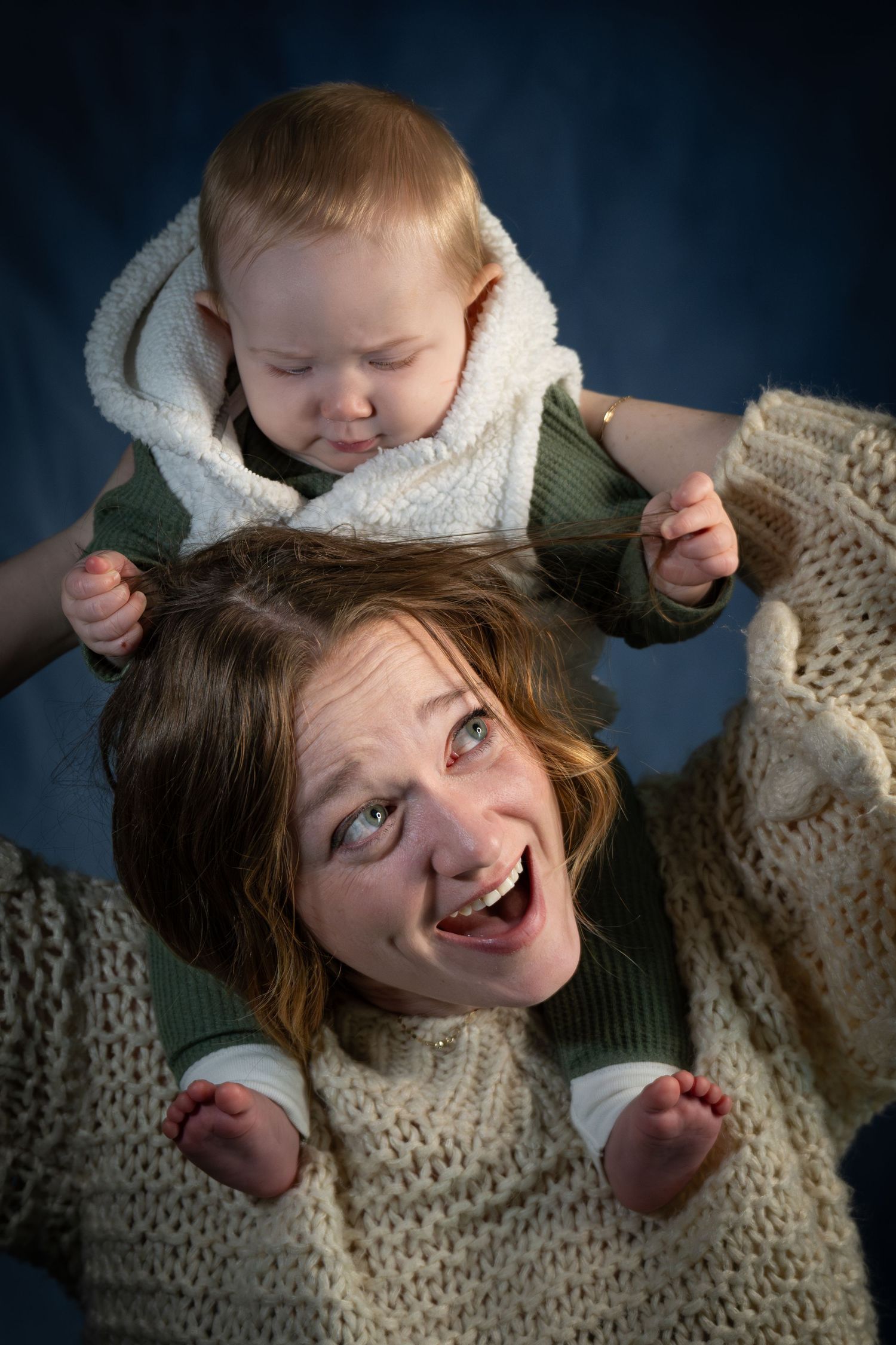 Laughing mother holds baby on her shoulders in matching cream knit sweaters against a dark blue background.