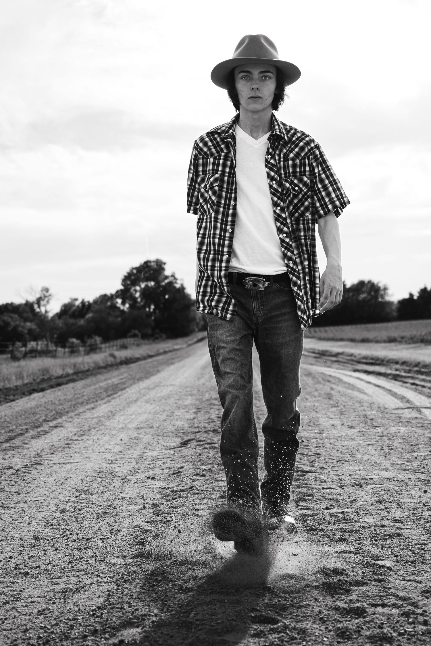 Black and white photo of a young man in a hat and plaid shirt walking on a rural dirt road.