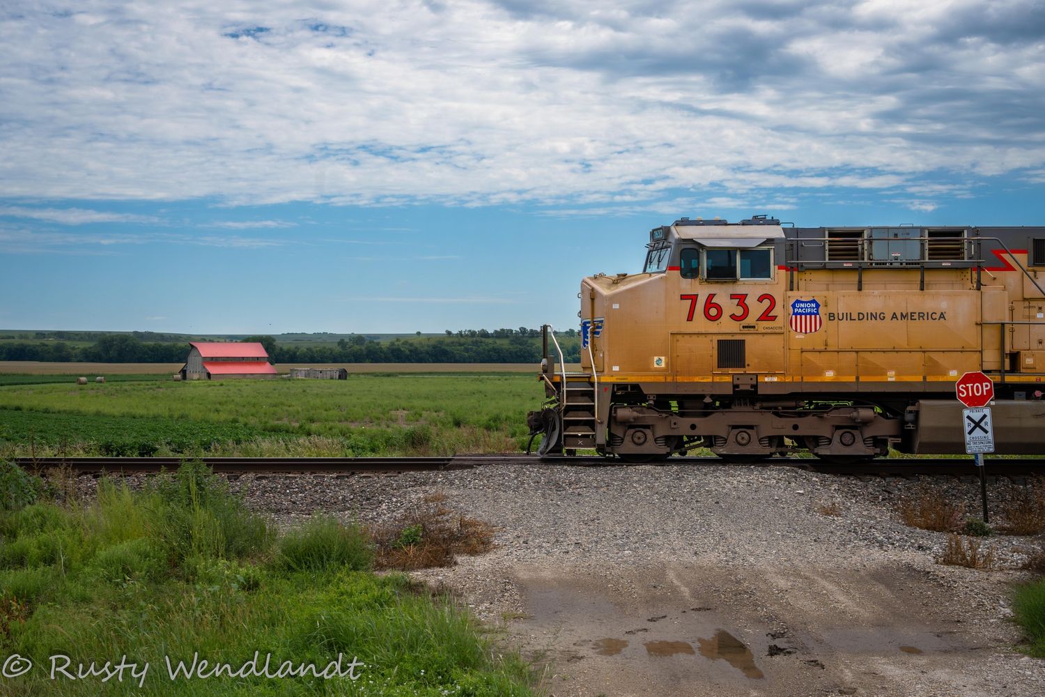 Long freight train moving through countryside with scattered buildings and cloudy sky. Union Pacific Kansas old barn