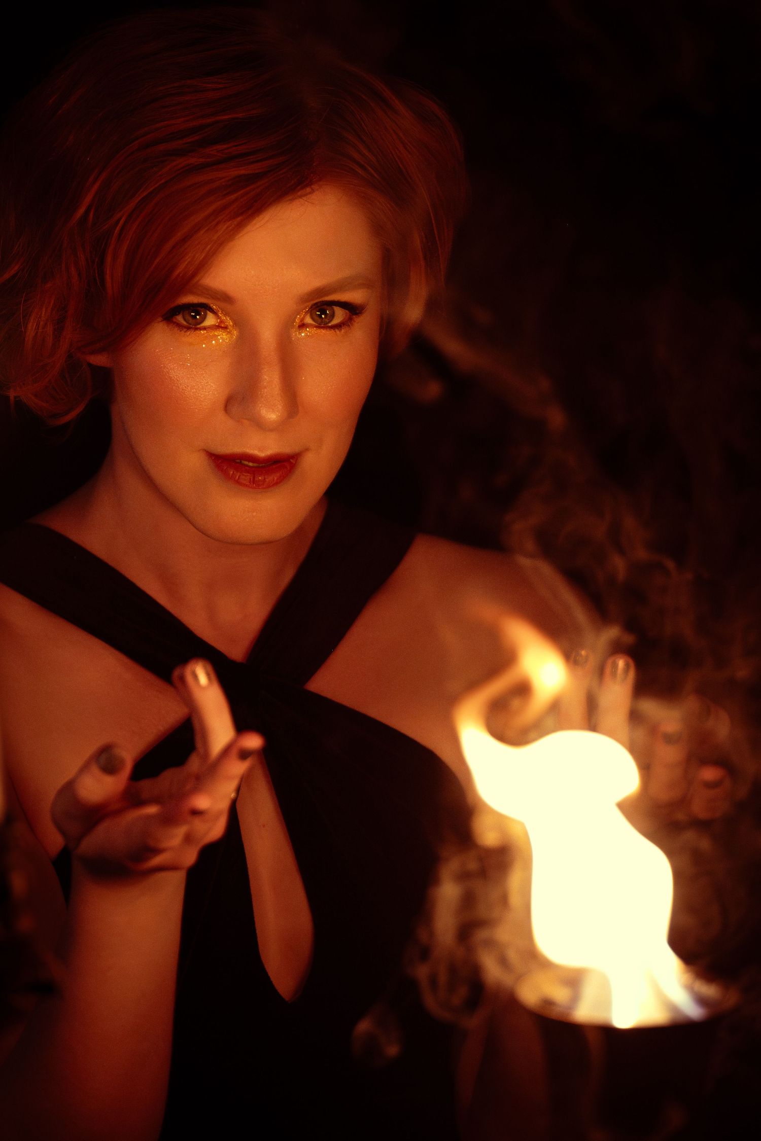 Red-haired woman in black dress holds a glowing flame in her hands against a dark background.