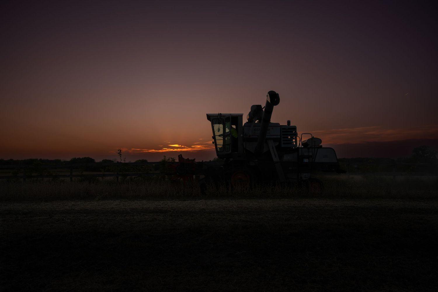 Dark silhouette of farm equipment against a dusky sunset horizon. Gleaner E combine harvesting soybeans sunset Wisconsin.