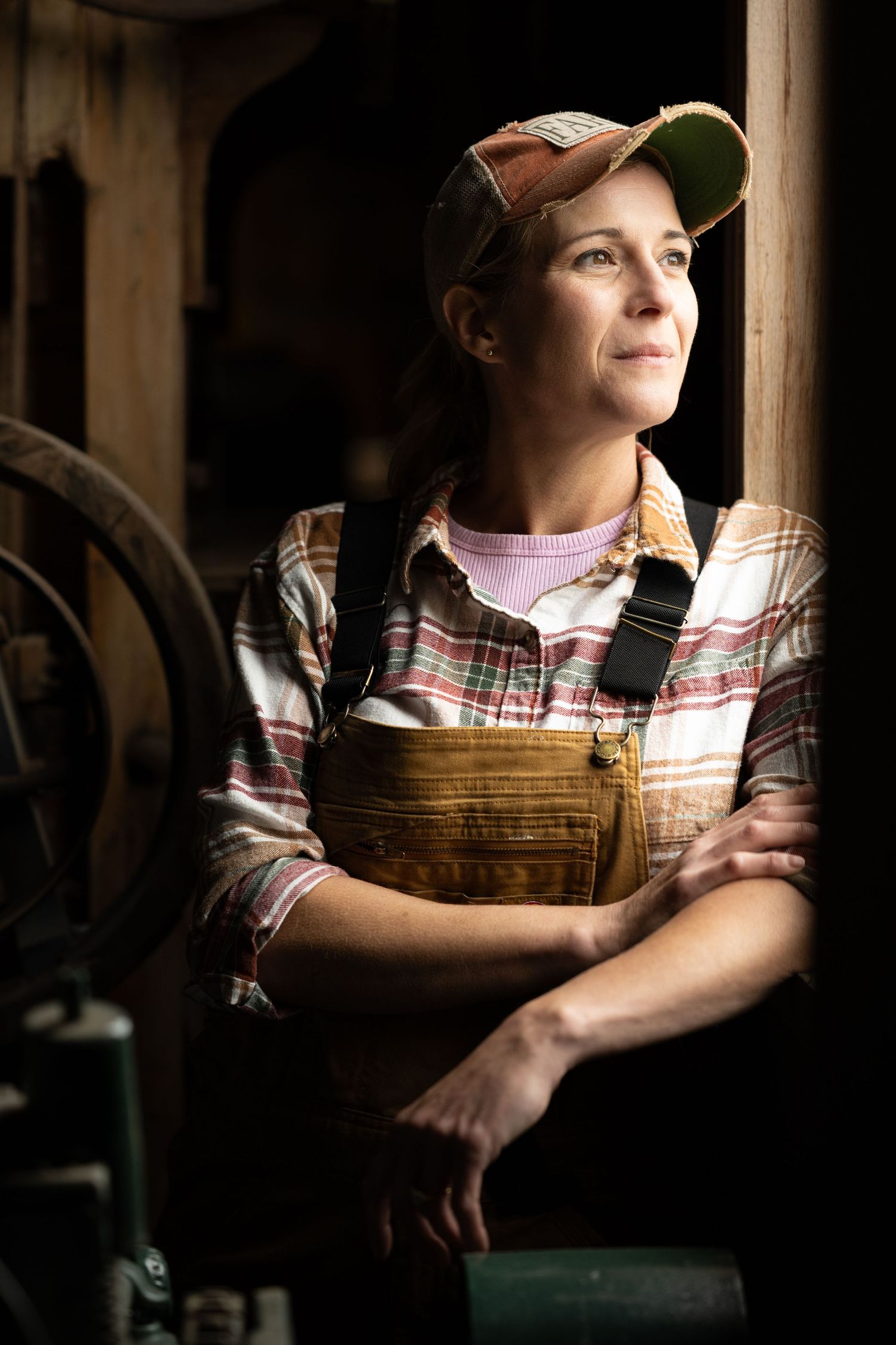 Female farmer in overalls and plaid shirt leans thoughtfully in rustic barn doorway.