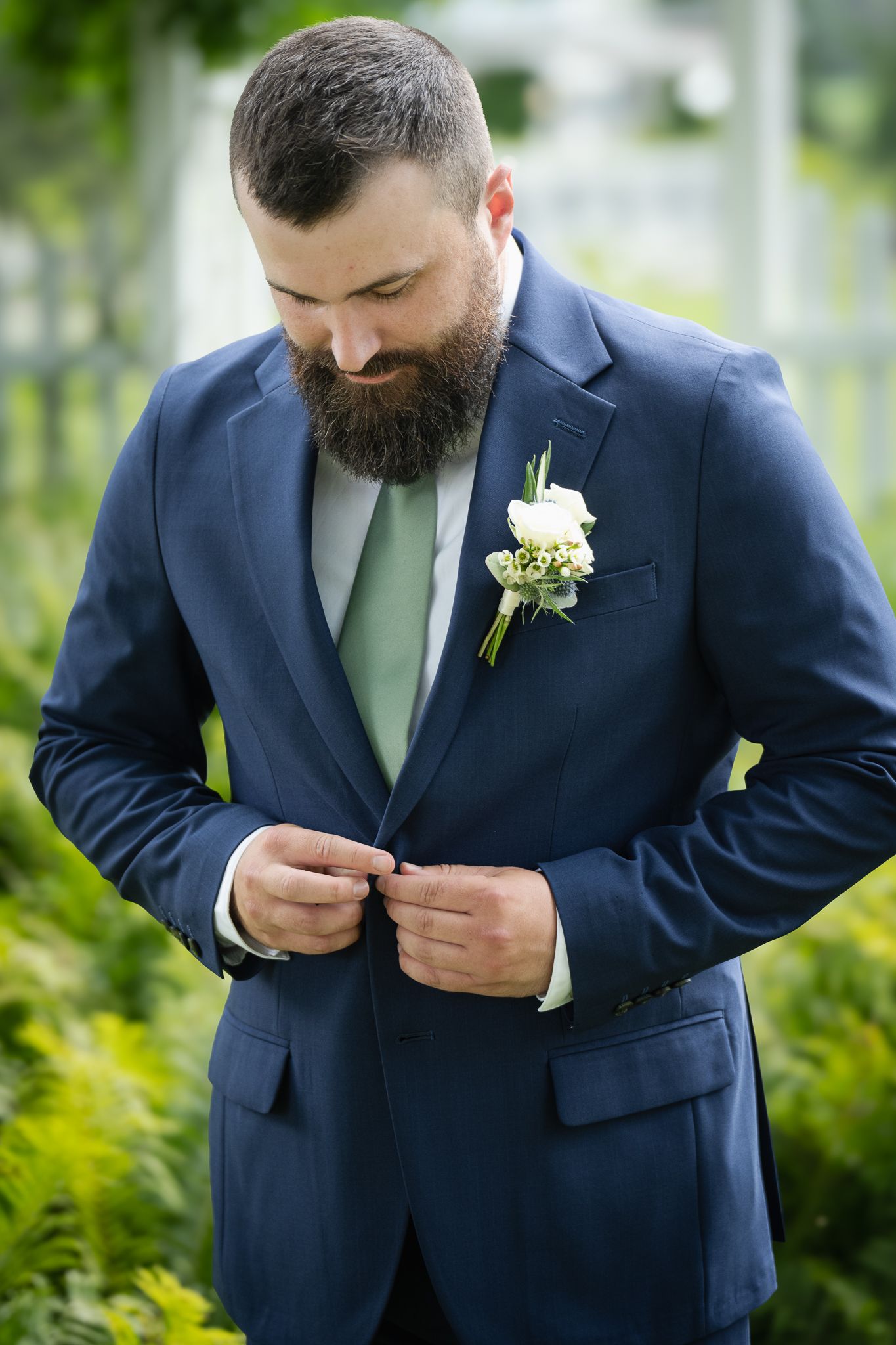 Bearded groom in navy suit with sage tie and white boutonniere buttons his jacket in lush green garden.