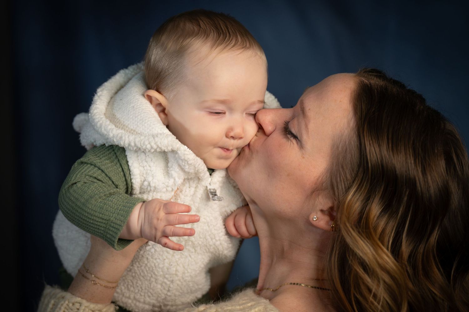 A mother tenderly kisses her smiling baby dressed in a white fluffy vest against a blue background.