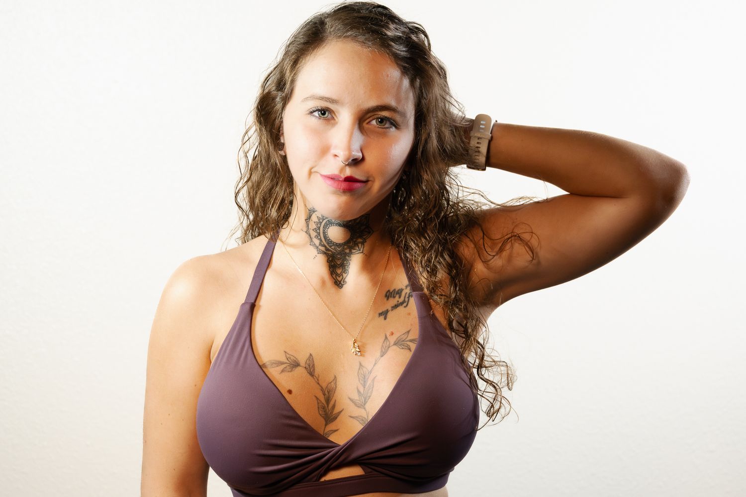 A young woman with tattoos and wavy hair poses in a brown bikini top against a white background.