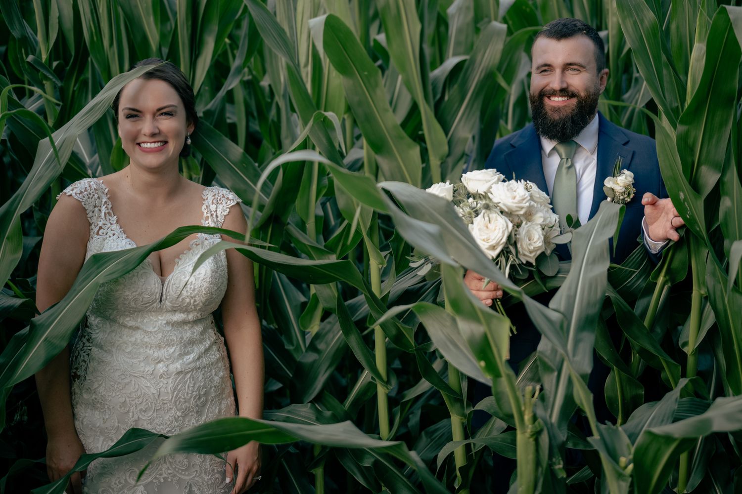 Bride and bearded groom smile among tall green cornfield holding white flower bouquet.