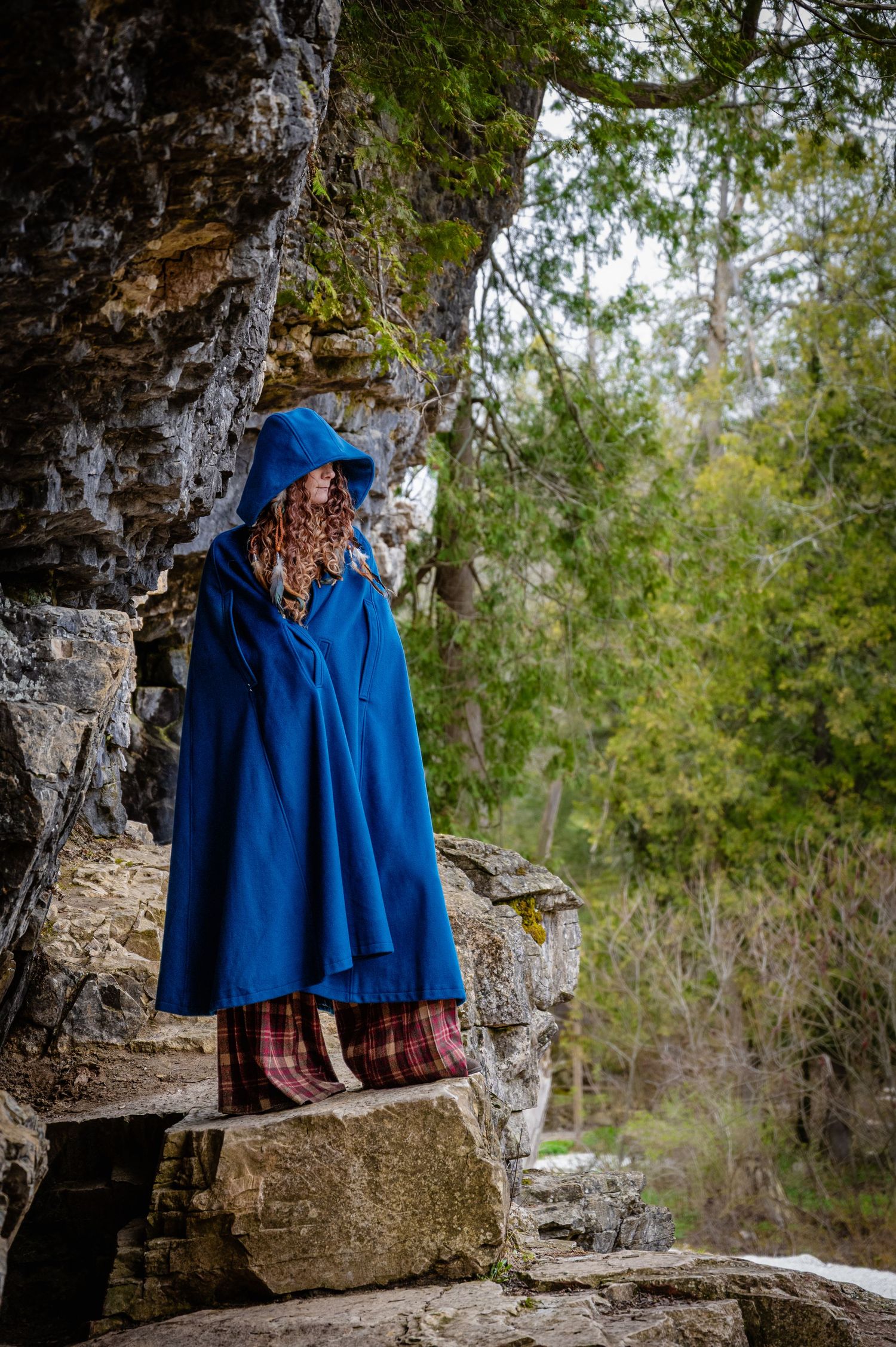 Person in a blue hooded cloak stands on stone steps surrounded by forest trees.