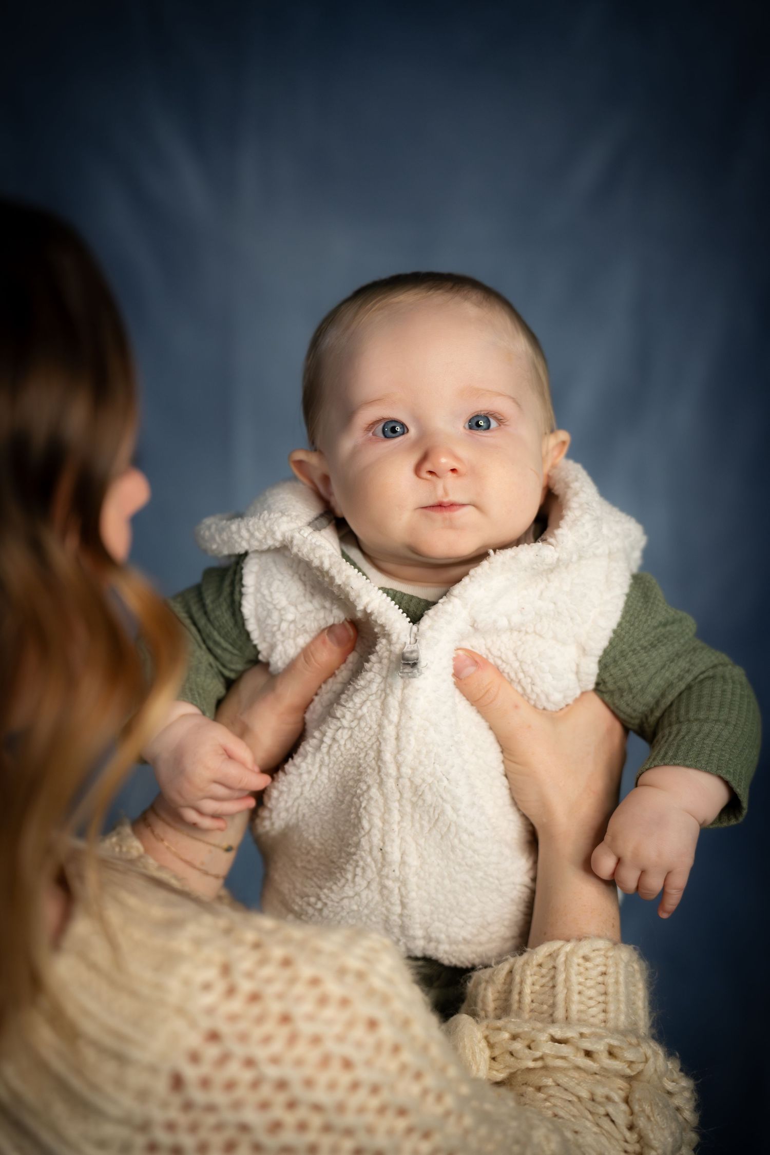 Mother in cream knit sweater holding adorable baby in white fluffy vest against blue backdrop.